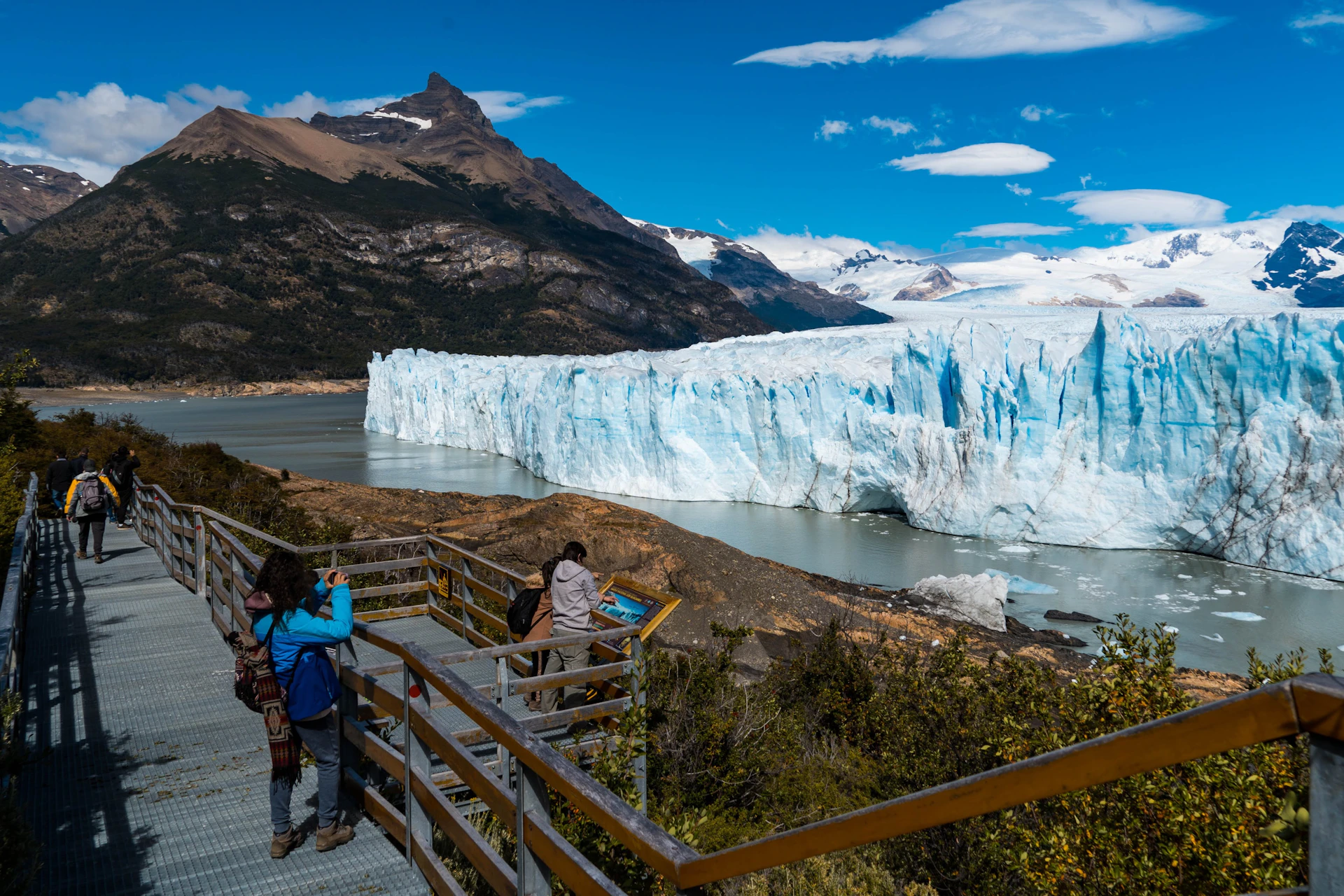 Tourists on a wooden walkway viewing a large glacier surrounded by mountains and water under a partly cloudy sky.