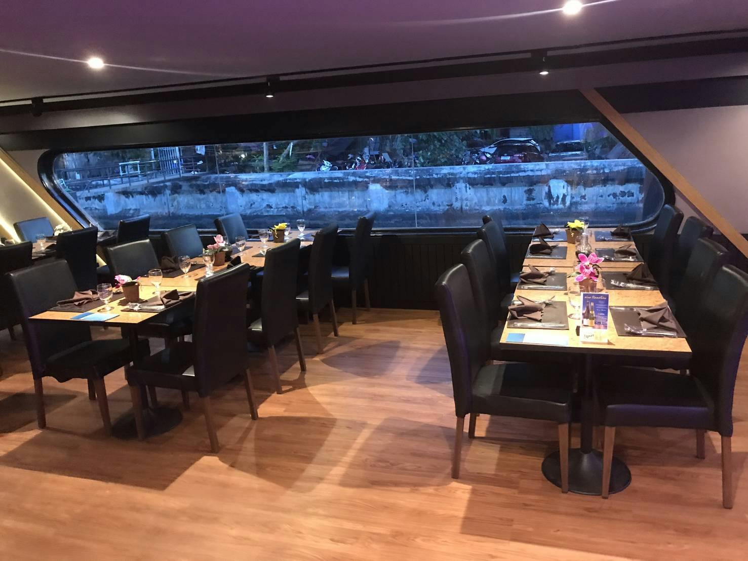 Jídelna s více stoly pro hosty; tmavé židle, dřevěná podlaha, velké okno s výhledem na městský kanál.