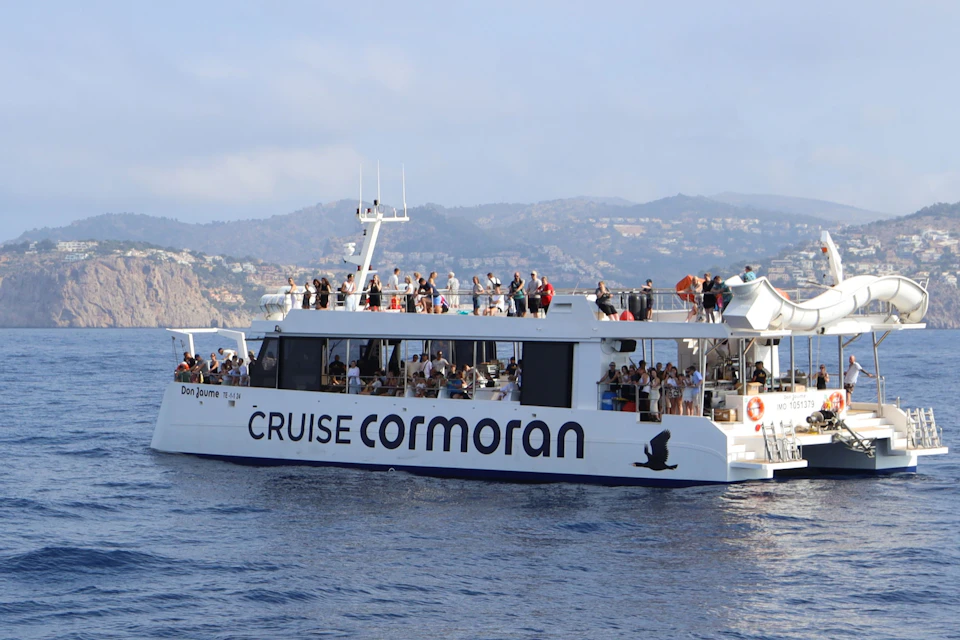 A white cruise boat named 'Cruise Cormoran' on water with passengers aboard, set against a coastal backdrop.