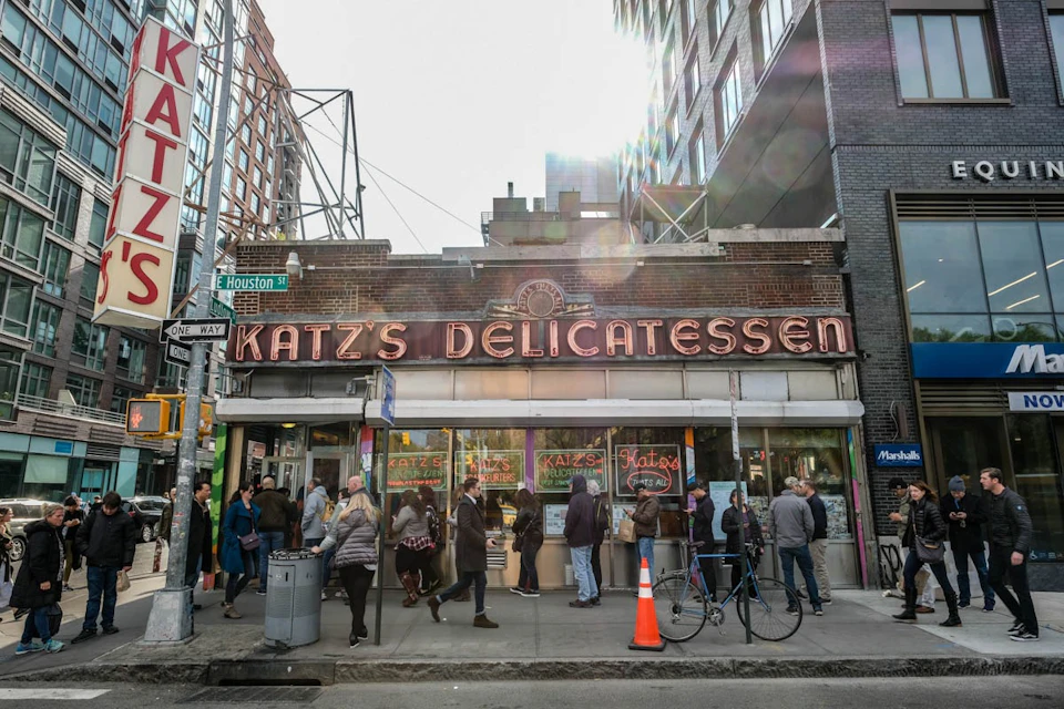 People lined up outside Katz's Delicatessen on a busy street with bicycles and a construction cone near the entrance.
