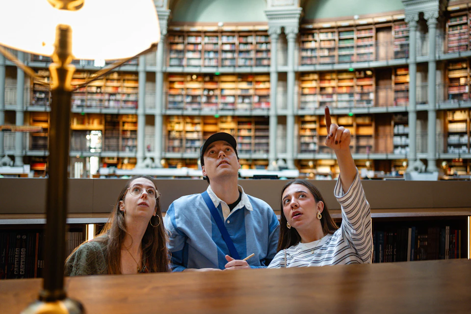 Three people sitting at a table in a library, looking upwards, with bookshelves and arched architecture in the background.