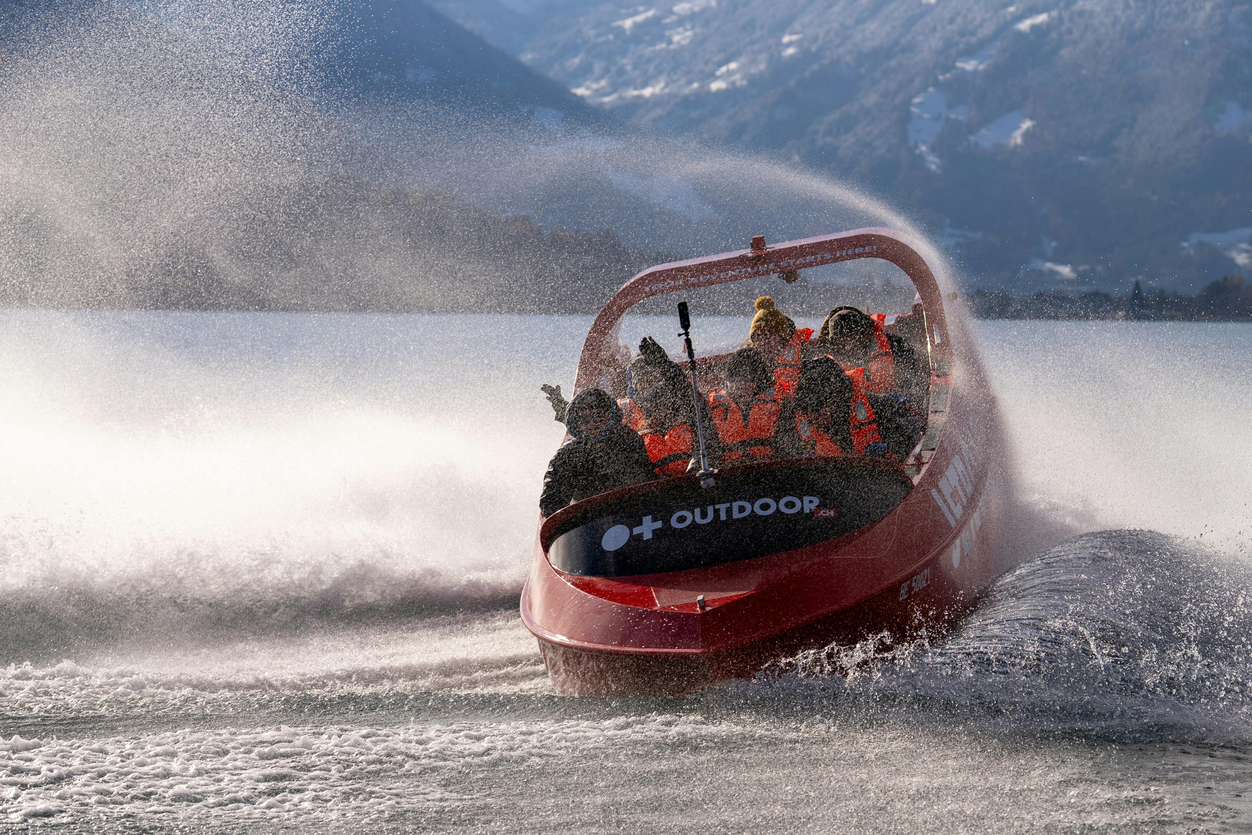 A red speedboat with passengers in life jackets moves through water, creating spray, with mountains visible in the background.