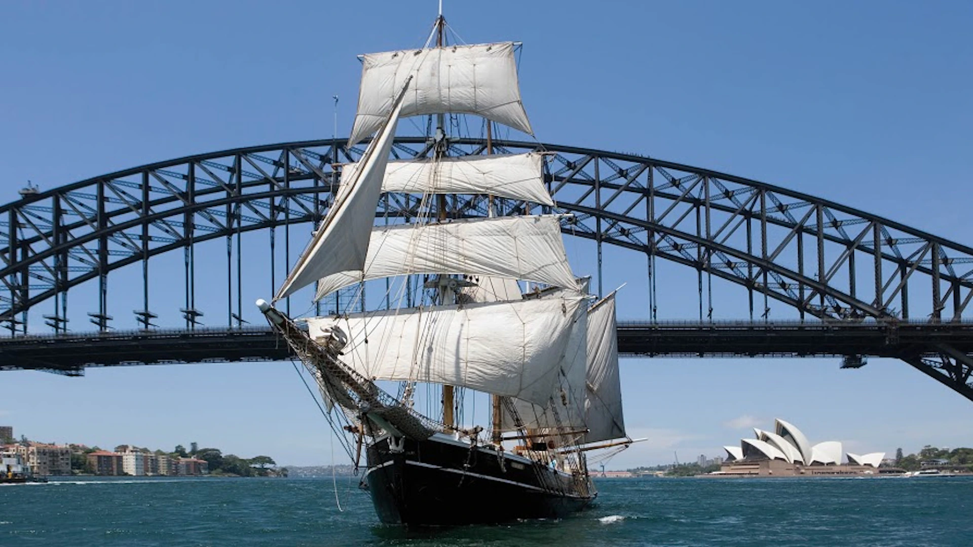 A sailing ship with white sails on the water, the Sydney Harbour Bridge, and the Sydney Opera House in the background.