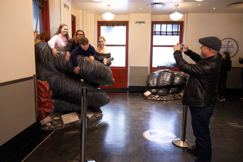 Four people pose on a large gorilla hand sculpture inside a room while another person takes their photo.