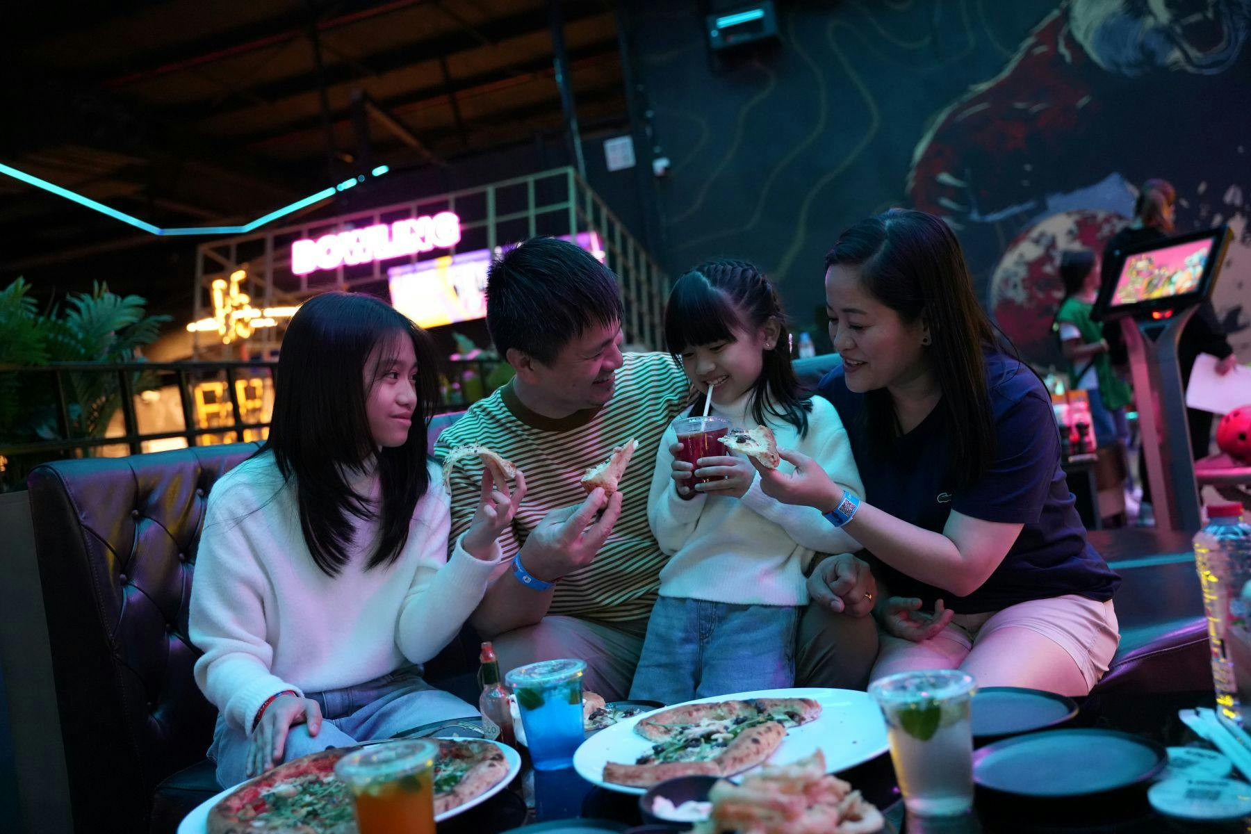 Μια οικογένεια που απολαμβάνει πίτσα και ποτά μαζί σε μια ζωντανή, πολύχρωμη τοποθεσία με θέμα τα arcade.