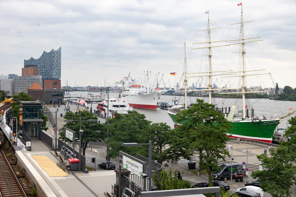 View of a busy harbor with ships docked, including a tall sailboat and large vessel. Pedestrians and buildings are visible near the waterfront.