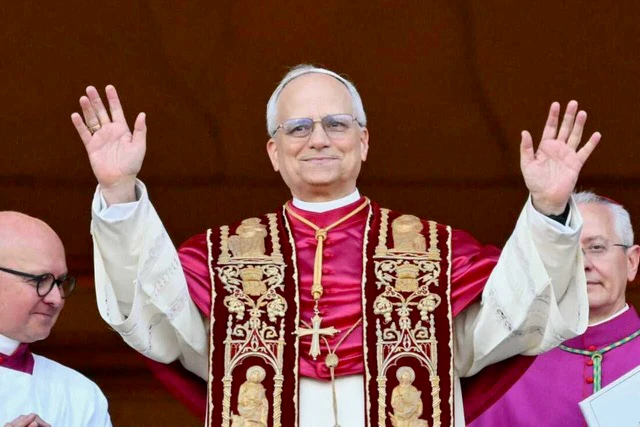 A man wearing religious garments and glasses smiles with his hands raised, standing against a dark background.