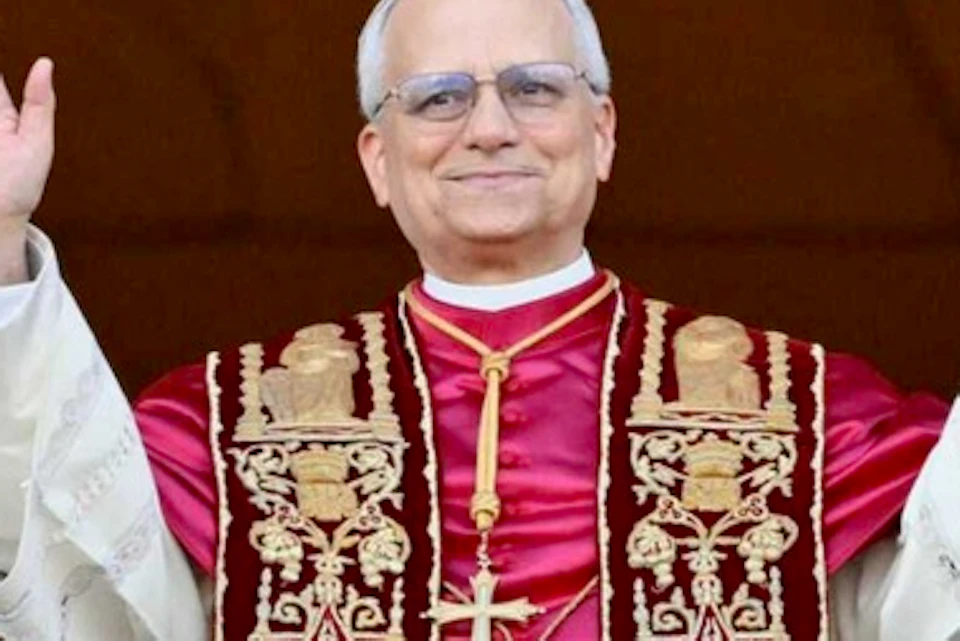 A man wearing religious garments and glasses smiles with his hands raised, standing against a dark background.