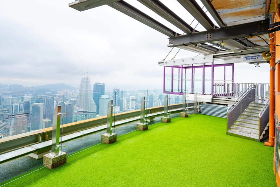 A rooftop terrace with green artificial grass, glass railings, and a cityscape view including tall buildings in the background.