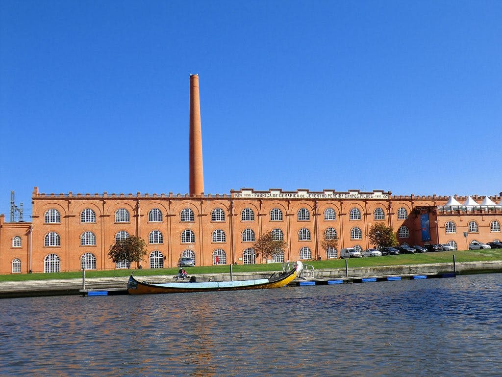 A brick building with arched windows and a tall chimney stands behind a river with a boat docked in the foreground under a clear blue sky.