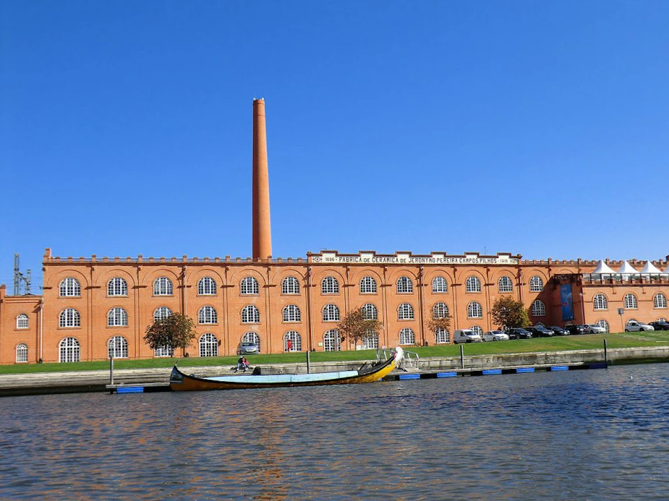 A brick building with arched windows and a tall chimney stands behind a river with a boat docked in the foreground under a clear blue sky.