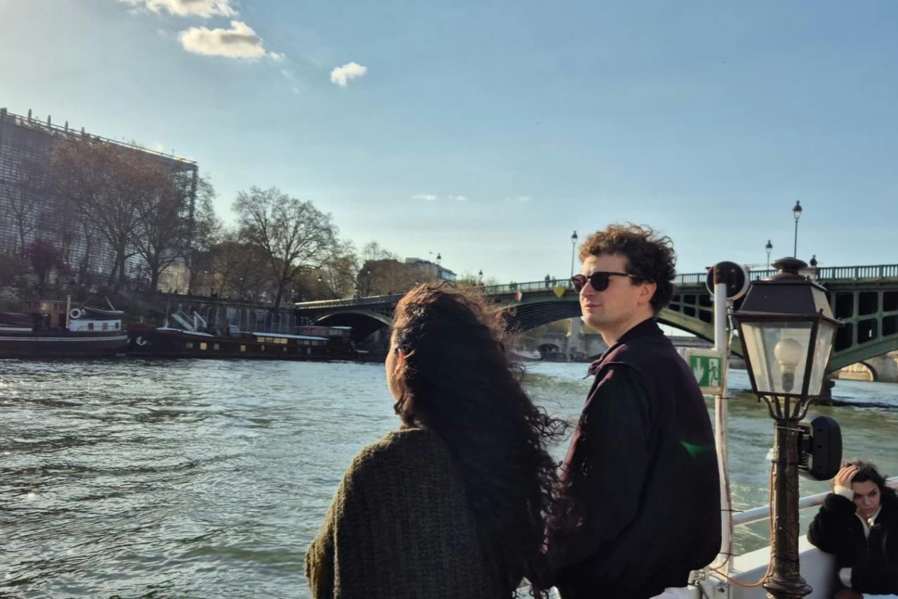 Two people stand on a boat near a river, with a bridge and lamppost visible in the background under a clear sky.
