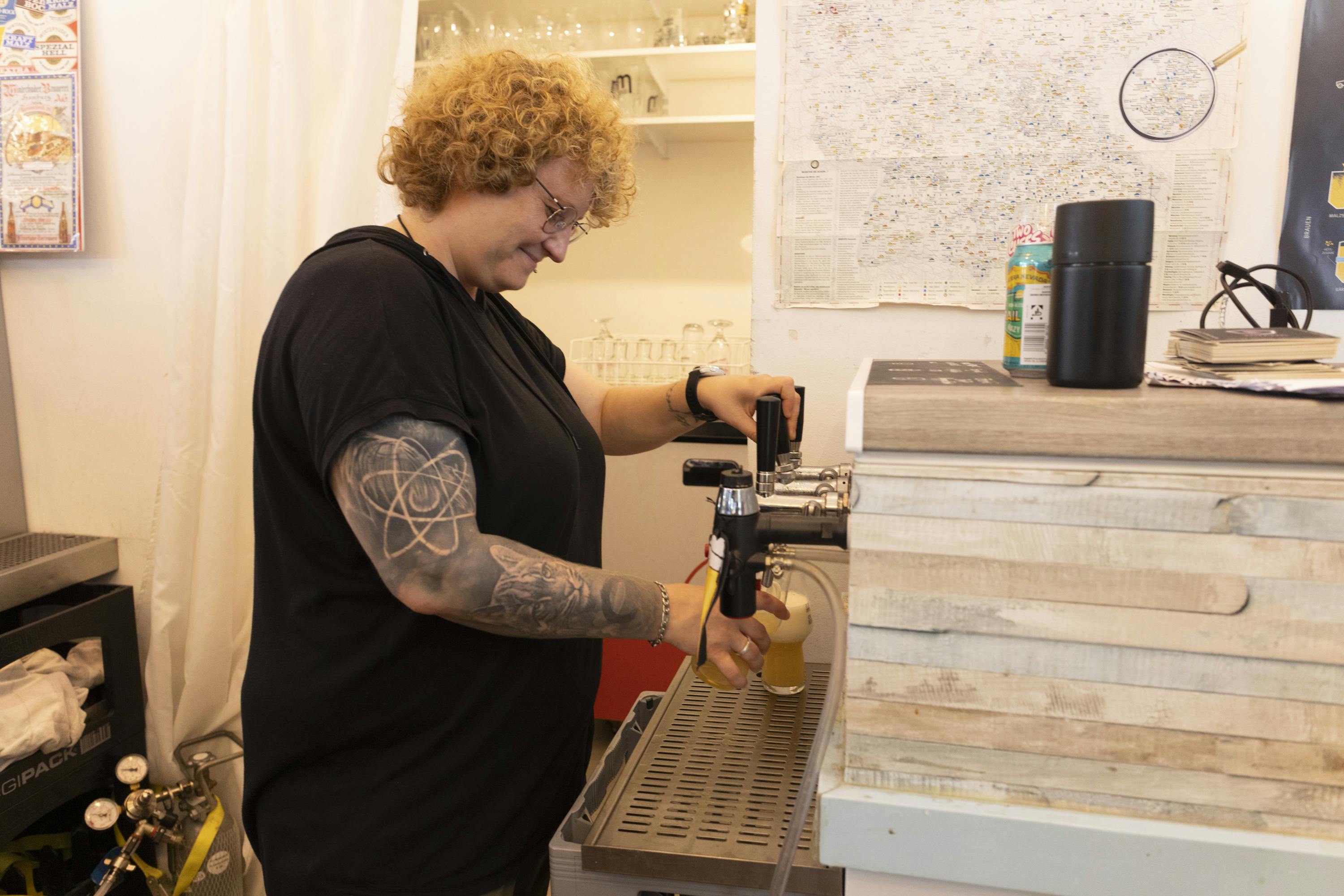 En person med krøllet hår og armtatoveringer hælder en drink fra en hane bag en bar. Hylder med glasvarer og et kort er i baggrunden.