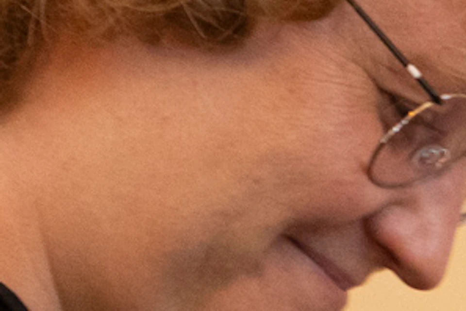Person with curly hair and arm tattoos is pouring a drink from a tap behind a bar. Shelves with glassware and a map are in the background.