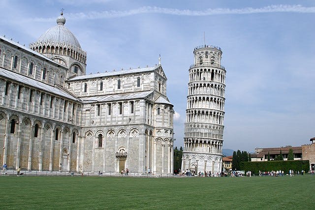 The Leaning Tower of Pisa and the Pisa Cathedral, with a clear sky and a green lawn in the foreground. Tourists are present.