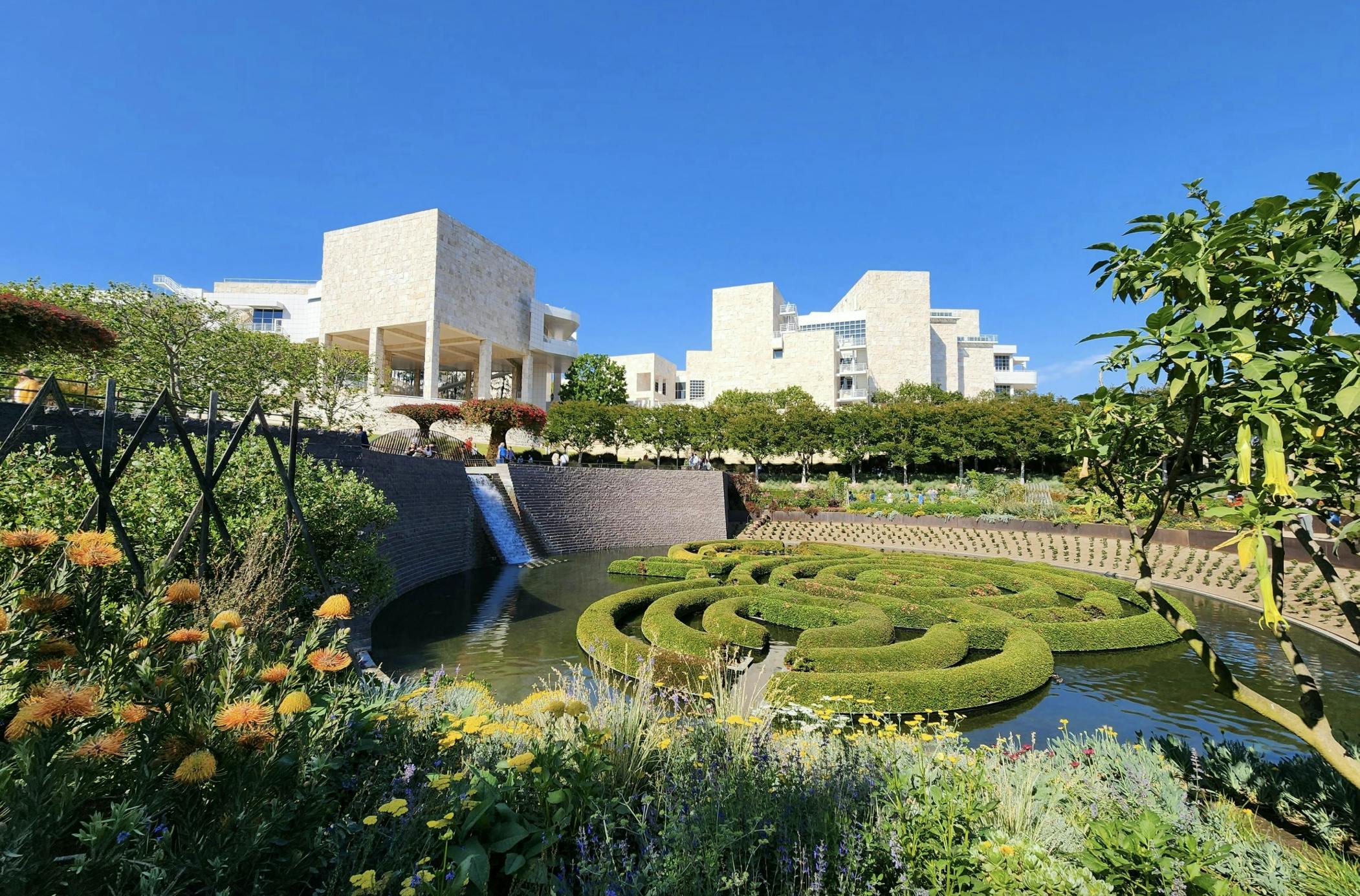 현대적인 석조 건물은 원형의 울타리 미로, 꽃, 나무, 맑고 푸른 하늘 아래 폭포가 있는 정원을 내려다보고 있습니다.