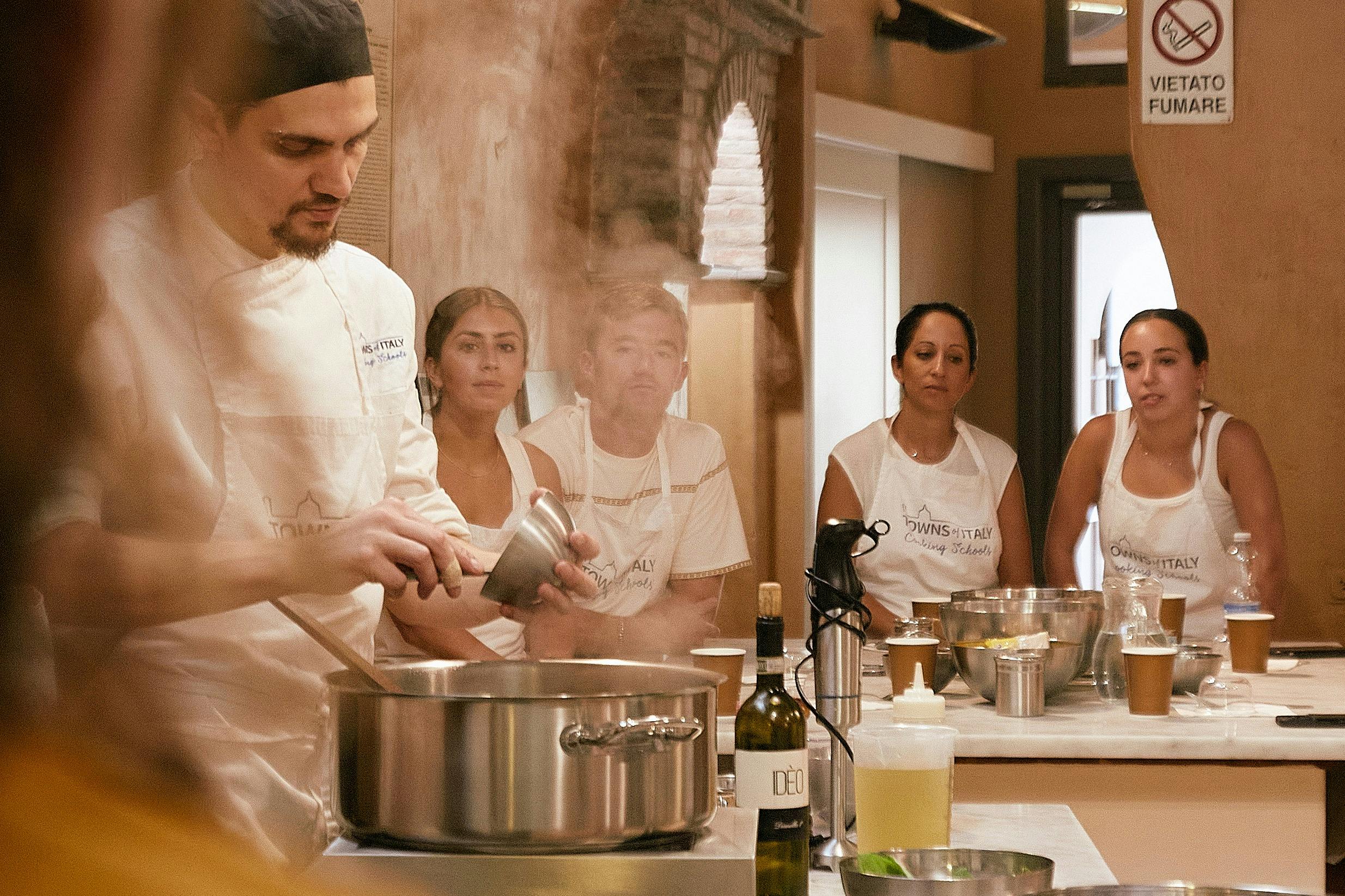 흰색 교복을 입은 셰프가 주방에서 요리하고 있으며, 네 명이 앞치마를 착용하고 있습니다. 조리도구와 식재료는 카운터에 있습니다.