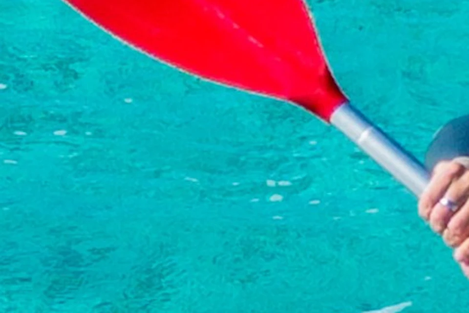 Two people in orange life vests kayaking on clear turquoise water, each holding a red paddle and smiling at the camera.