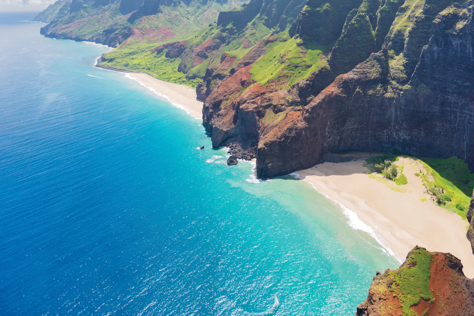 Aerial view of a coastline with steep green cliffs, a sandy beach, and clear blue ocean water.