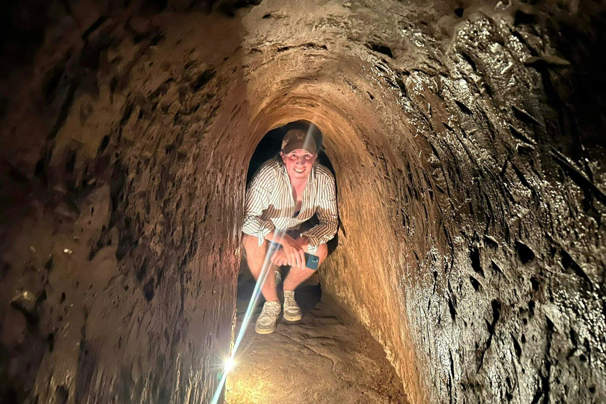 Person crouching in a narrow, dimly lit tunnel with textured walls, holding a smartphone and wearing a cap and striped shirt.