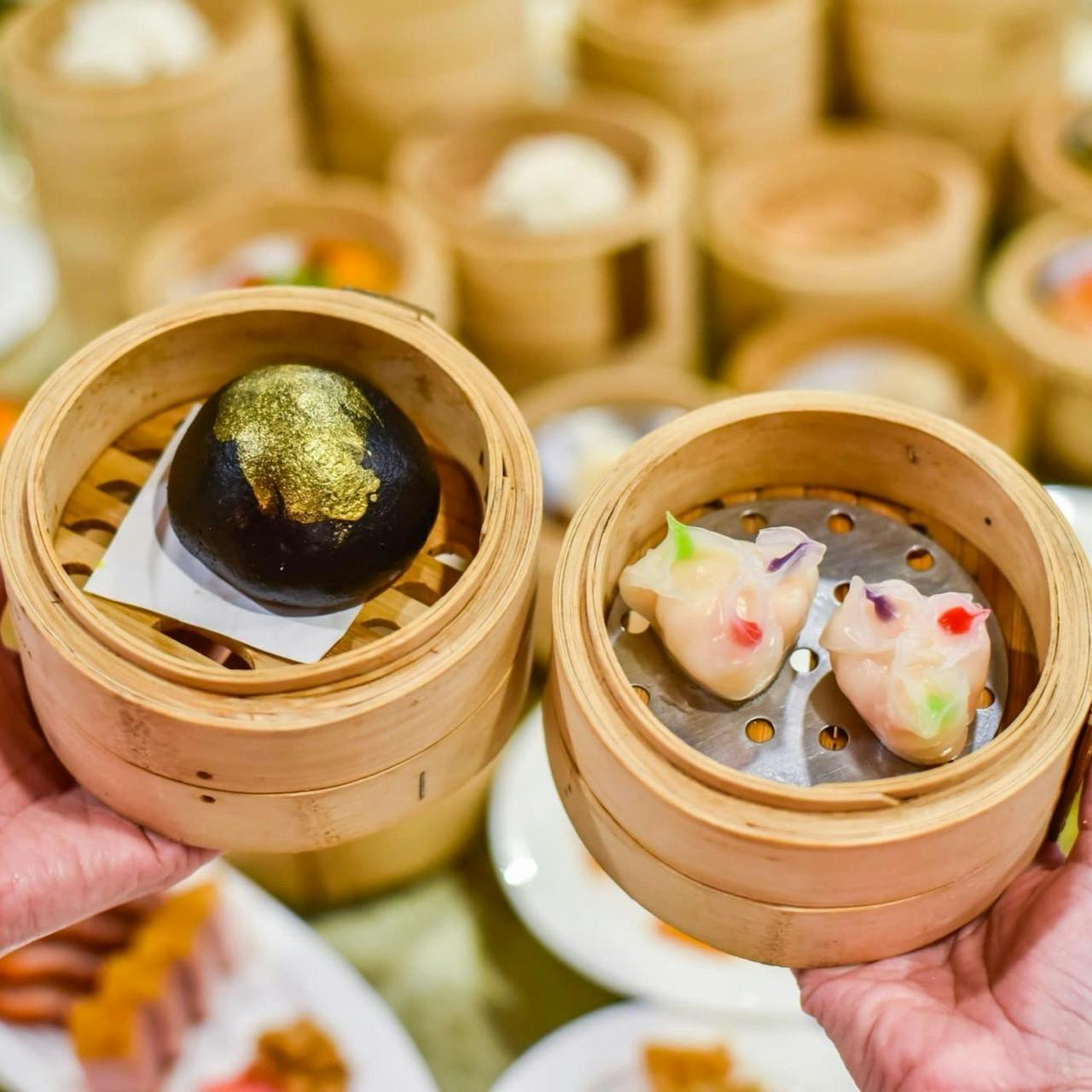 Hands holding bamboo steamers with a black bun topped with gold leaf and colorful dumplings, surrounded by more steamers.