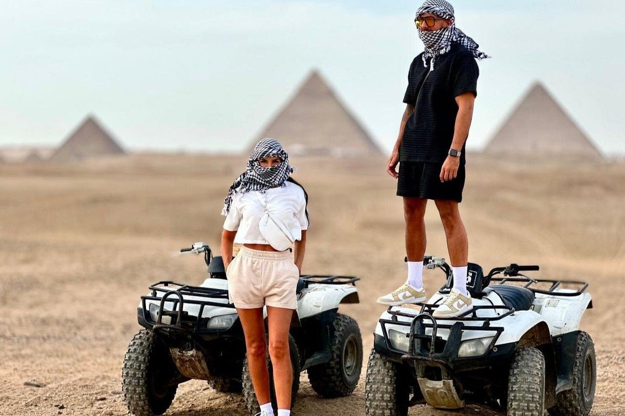 砂漠では、ピラミッドを背景に、ケフィエフに乗った2人の人物がATVと共に立っています。 片方はATVに乗っていて、もう片方はその横に立っています。