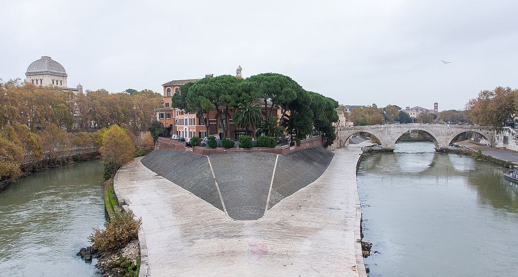 Island in a river with trees, buildings, and a bridge with multiple arches, surrounded by water and autumn trees. Cloudy sky.