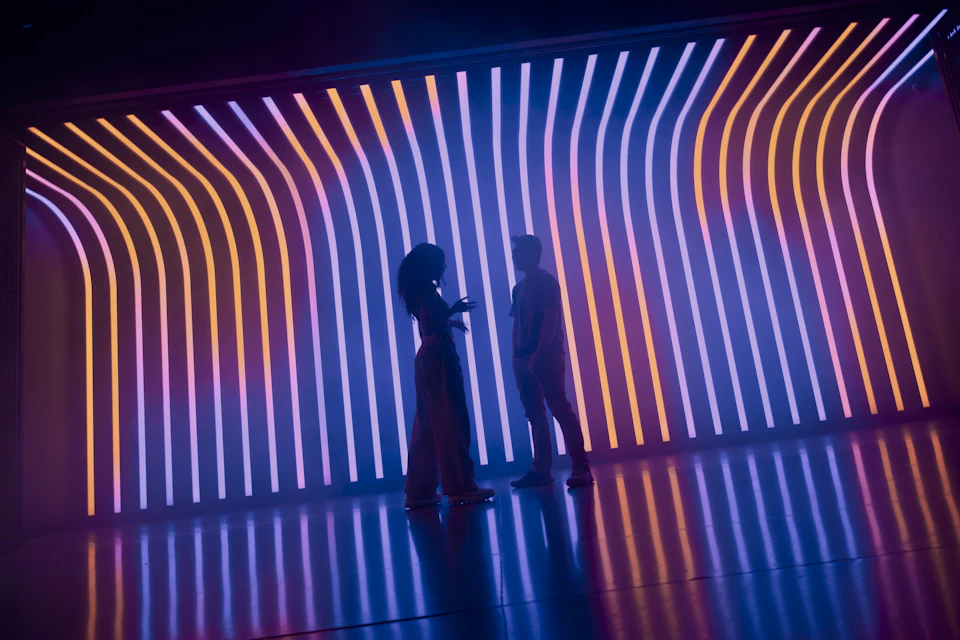 Two people stand silhouetted in front of a glowing wall with colorful, wavy vertical lines in a dimly lit room.