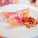 A white plate with thinly sliced prosciutto over melon and a piece of toast topped with diced tomatoes.