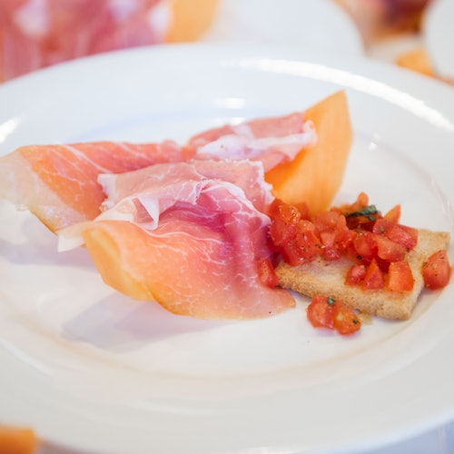 A white plate with thinly sliced prosciutto over melon and a piece of toast topped with diced tomatoes.
