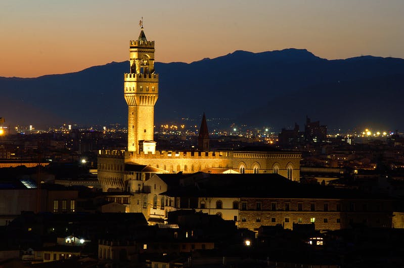 Una torre medievale illuminata al tramonto, con le luci della città e una catena montuosa sullo sfondo.
