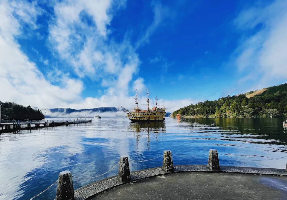 A scenic view of a lake with a ship resembling a pirate vessel, surrounded by mountains and a partly cloudy sky.