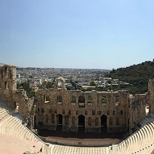 Forntida amfiteater i sten med nivåindelade sittplatser, delmurar och en avlägsen stadsbild under en klarblå himmel.