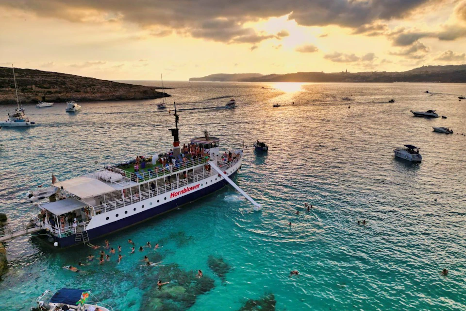 A boat named 'Hornblower' in a turquoise sea near swimmers, with a sunset and distant land visible in the background.