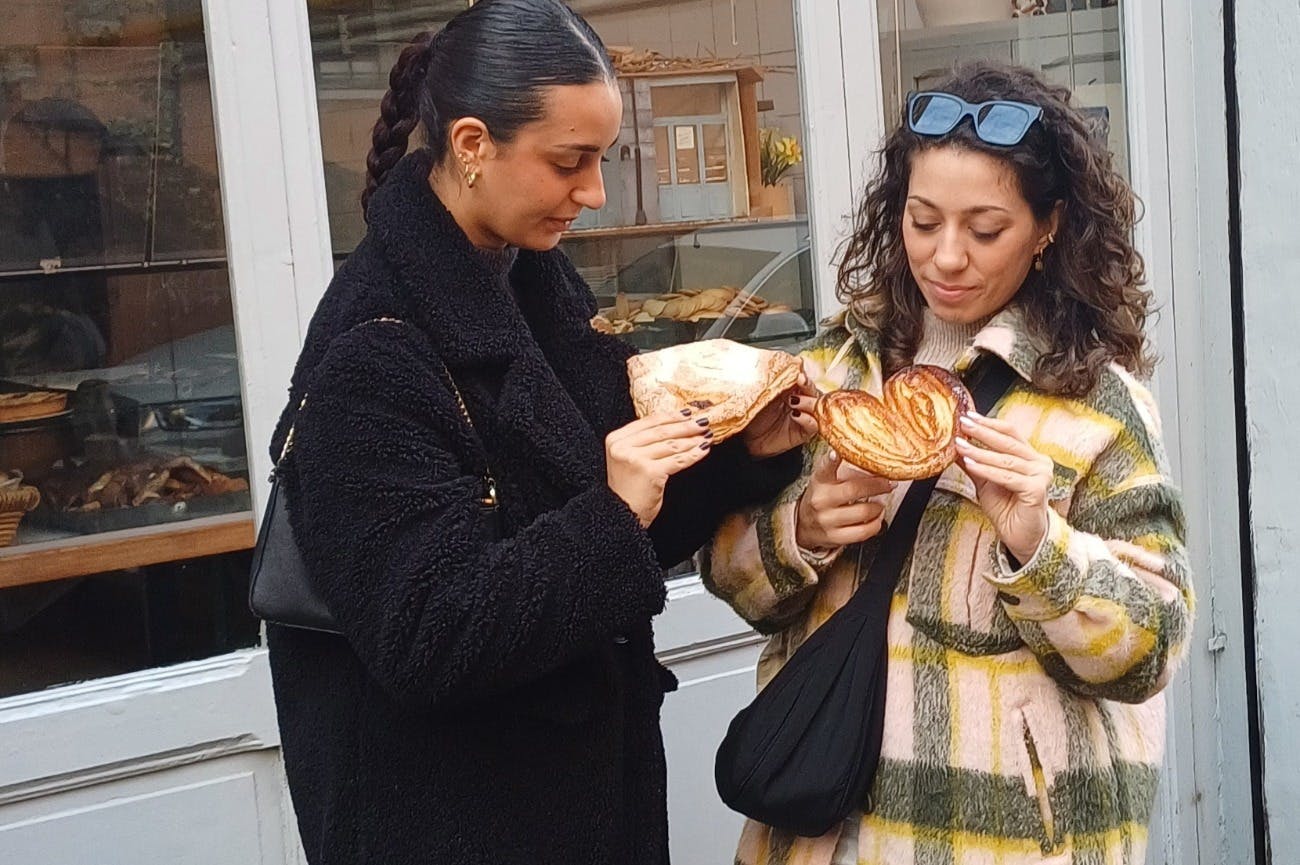 Two women in coats are holding and looking at pastries outside a bakery.