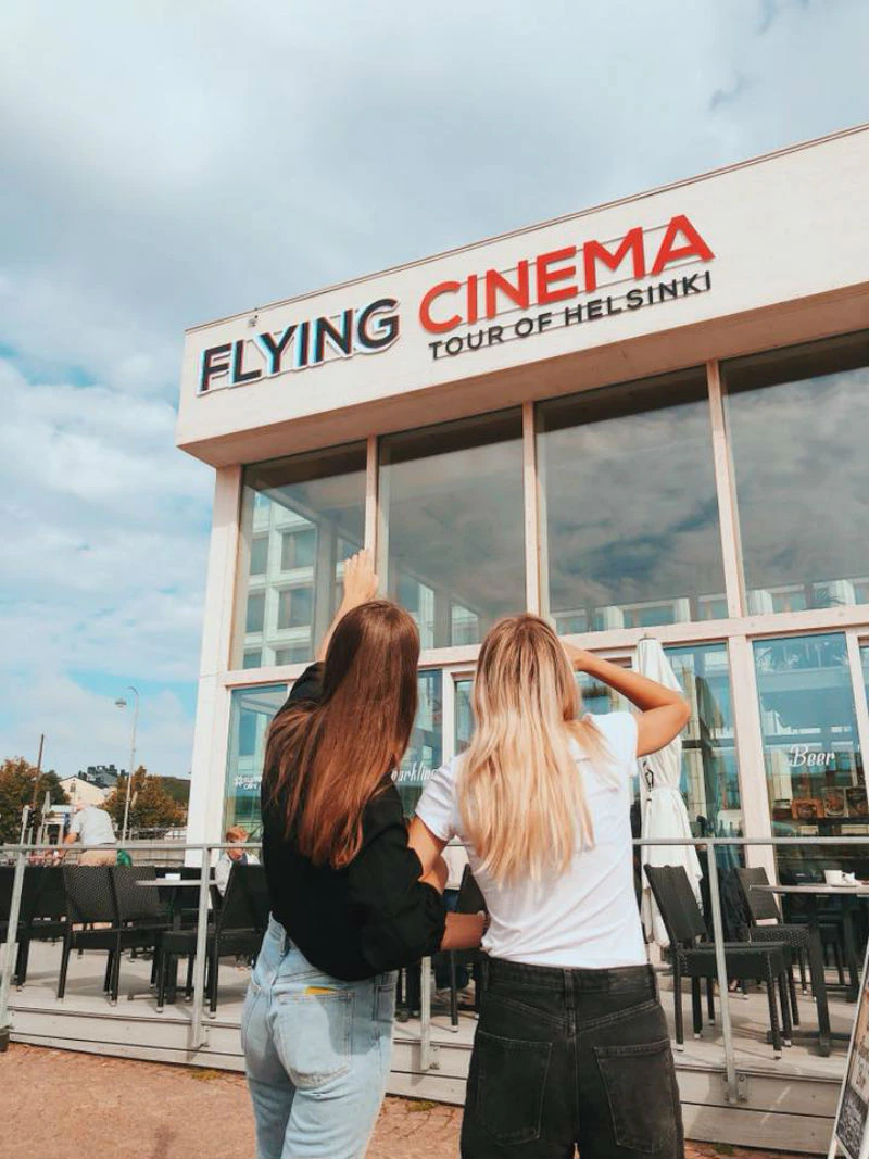 Two women with long hair stand outside a building labeled "Flying Cinema Tour of Helsinki,” looking up and pointing at the sign.