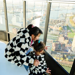 En kvinna och ett barn i matchande rutiga jackor tittar ut från ett högt observationsdäck vid en stadsbild med vattendrag och byggnader.
