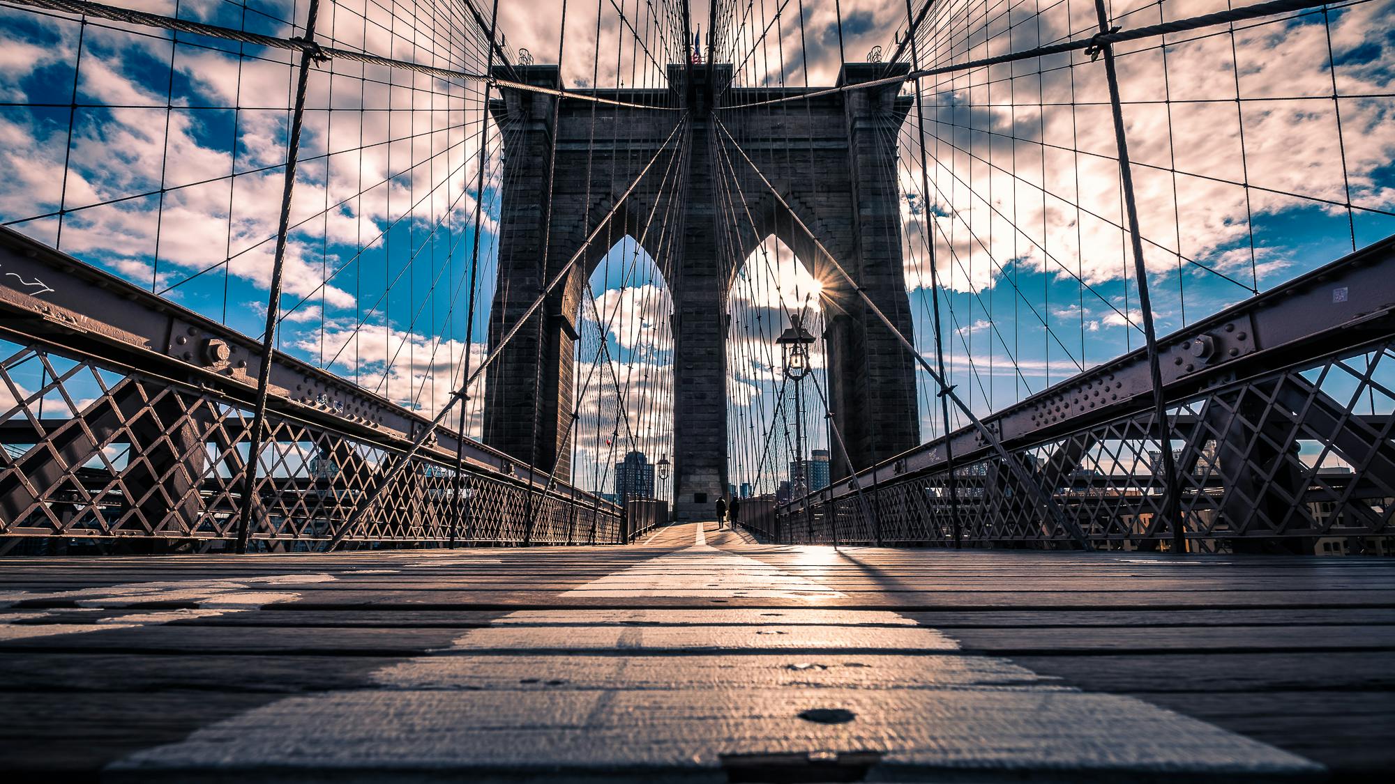 Una vista dal basso del ponte di Brooklyn con un'esplosione di sole attraverso i cavi, i dettagli architettonici e un cielo parzialmente nuvoloso.