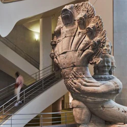 En stor stenskulptur av en flerhövdad orm med en mänsklig figur i en museumsmiljö, med en trappa i bakgrunden.