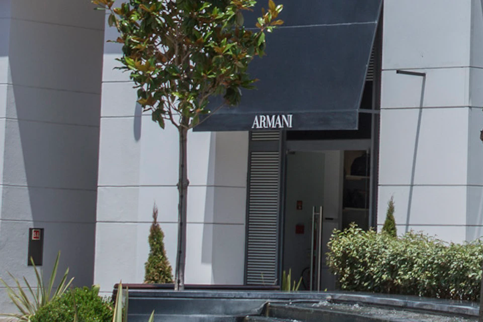 Three people in an outdoor shopping center with Giorgio Armani store. Two sit on a bench under a tree, while one walks carrying shopping bags.