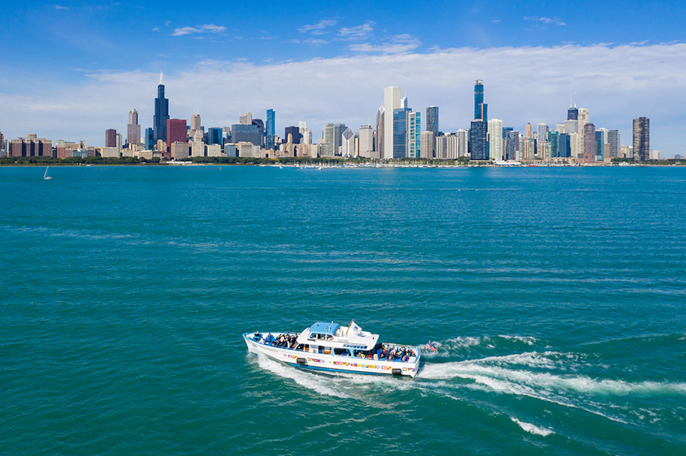 A tour boat with passengers sails on a large body of water, with a city's skyline and skyscrapers visible in the background.
