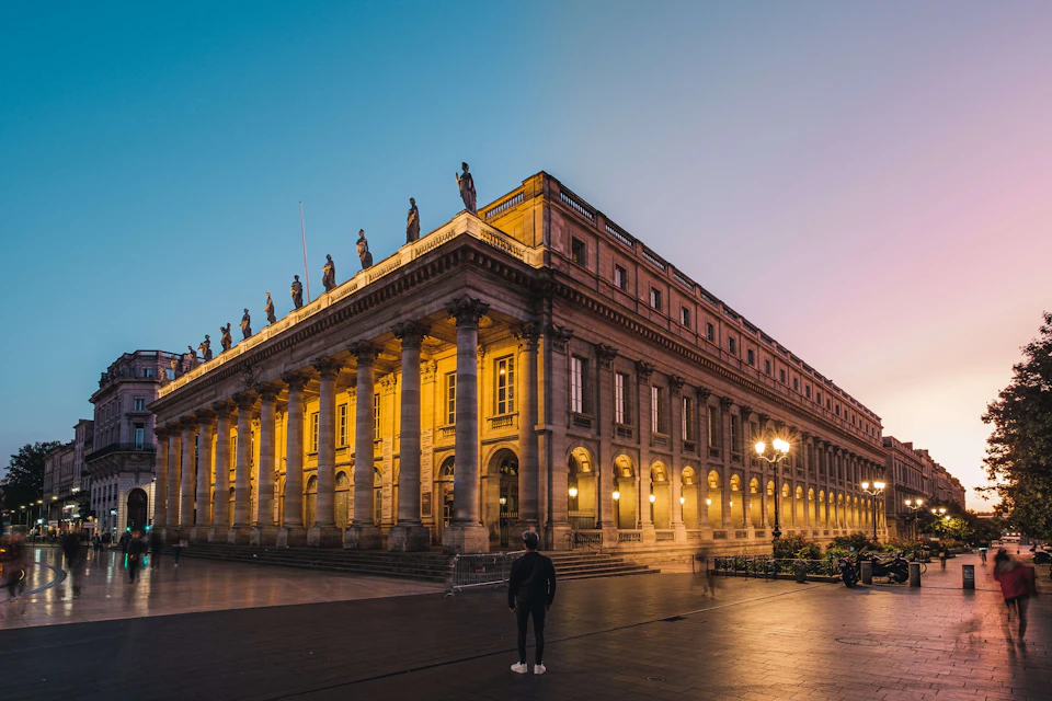 A person stands in front of an illuminated neoclassical building with columns and statues at dusk.