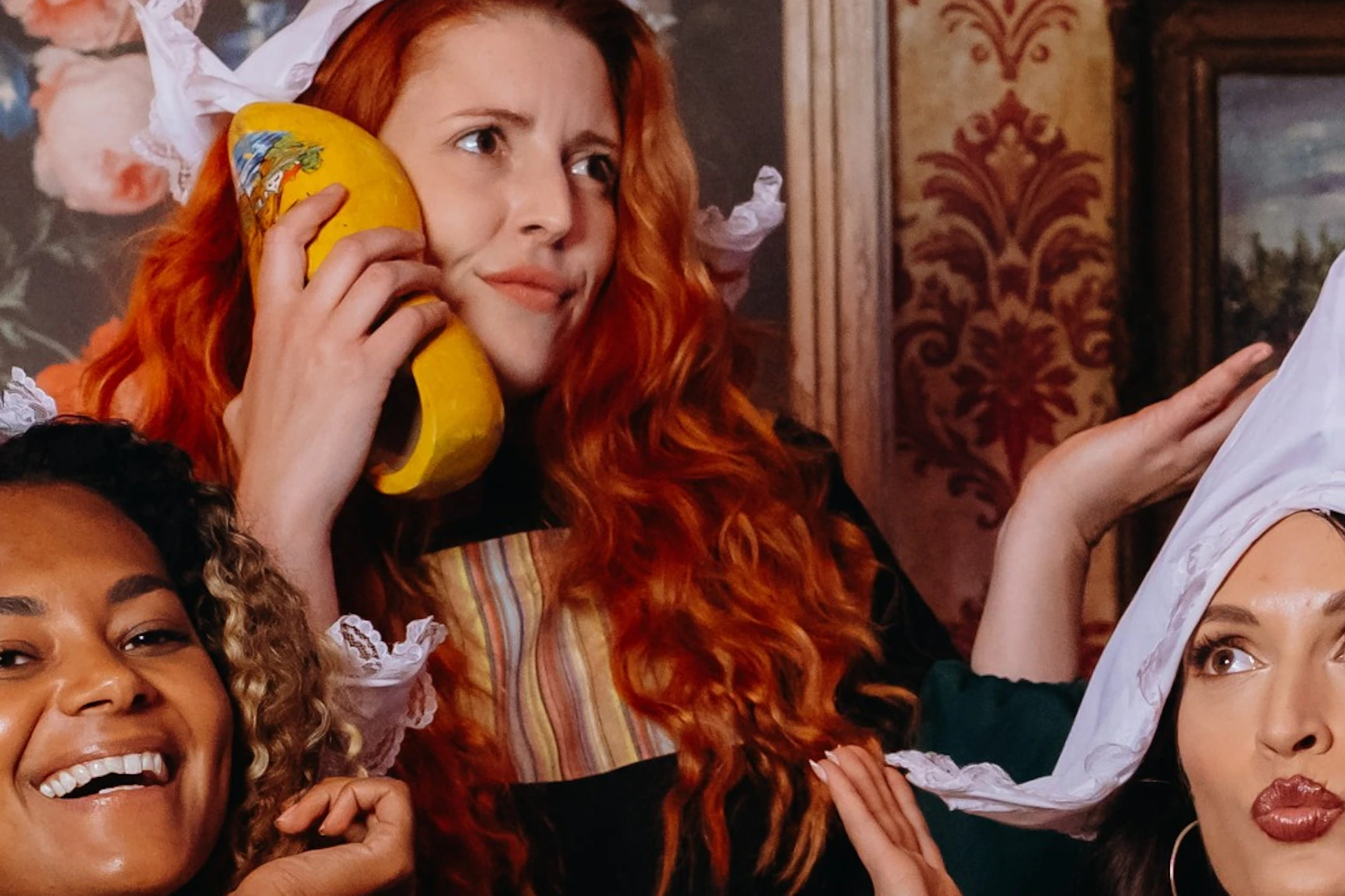 Three women in traditional attire holding wheels of cheese and tulips, posing in a vintage-themed room with floral wallpaper.