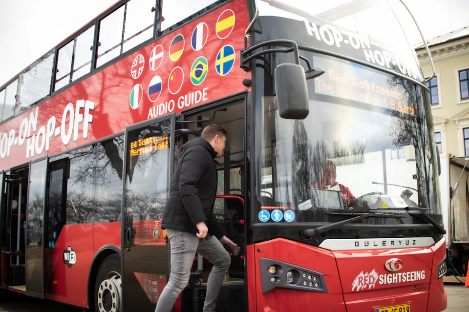 A person boarding a red double-decker sightseeing bus with flags and "Audio Guide" written on the side.