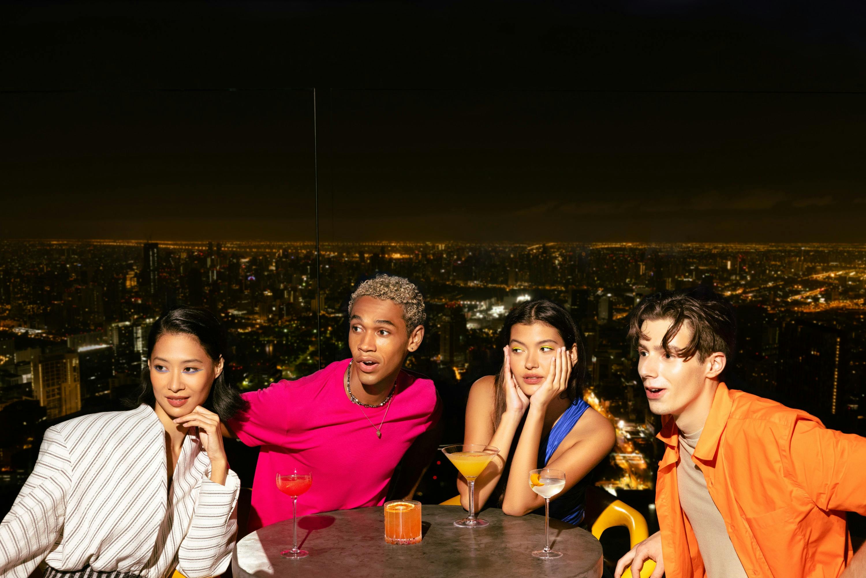 Four people seated around a table with cocktails, overlooking a cityscape at night.