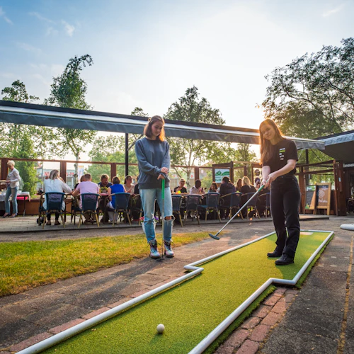 Två personer spelar minigolf utomhus nära en sittgrupp med flera personer som äter. Träd och en staty syns i bakgrunden.
