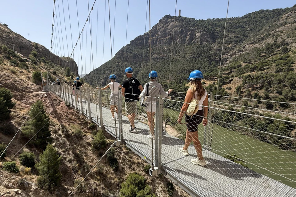 People wearing helmets walk on a suspension bridge over a scenic valley with hills and vegetation under a clear sky.