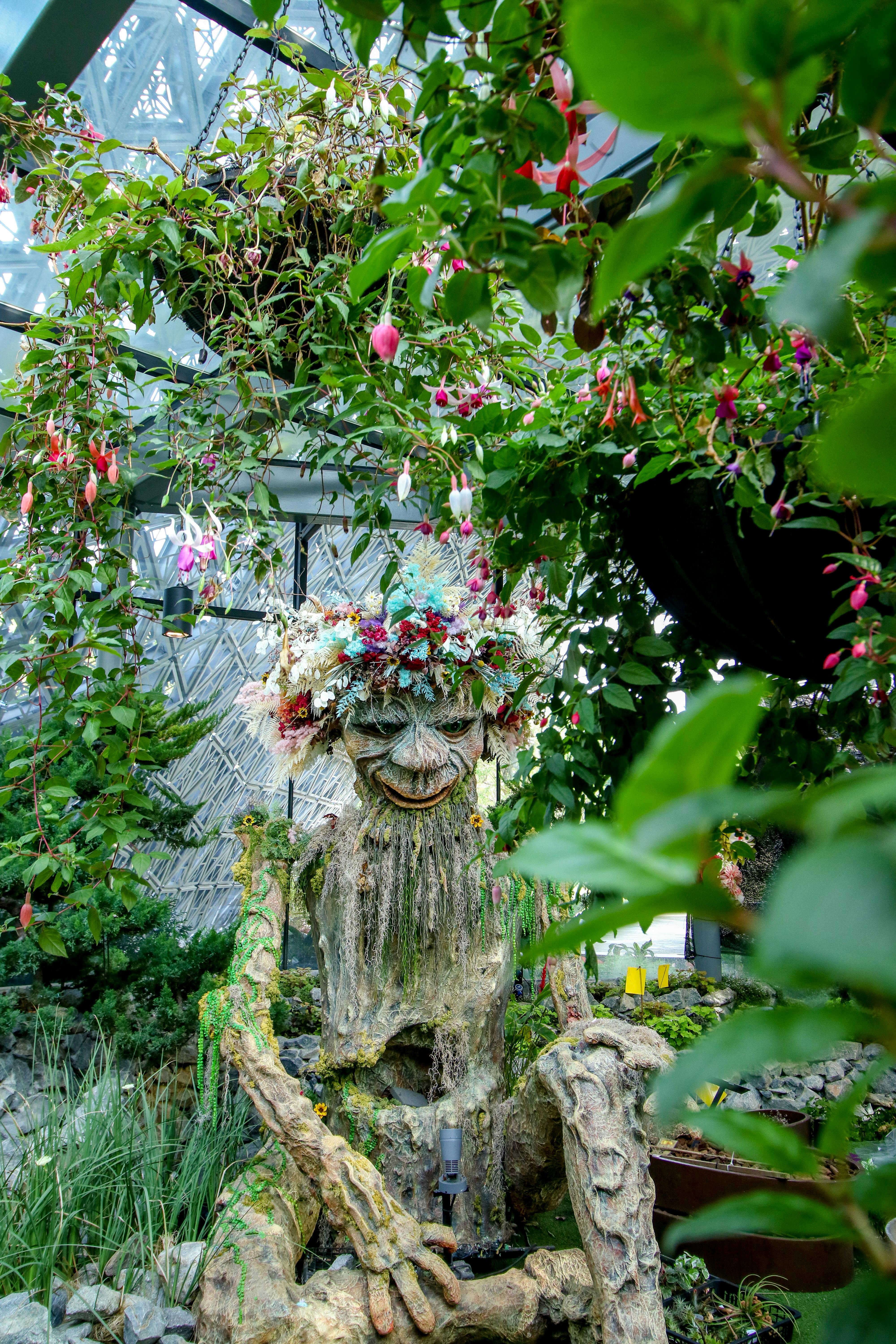 A tree-like sculpture with a face and flower crown surrounded by lush foliage and hanging flowers inside a greenhouse.