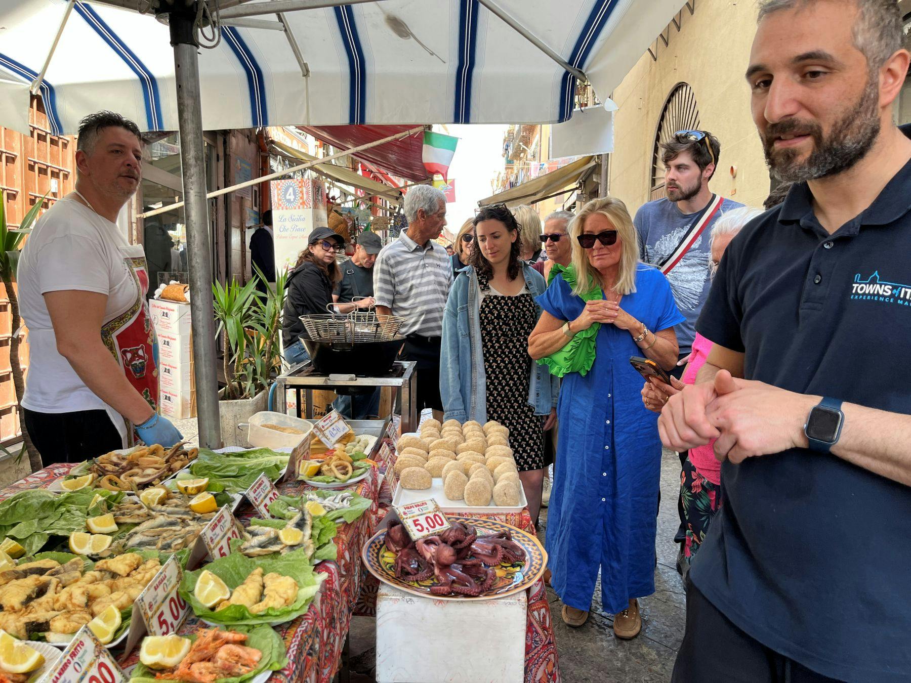 People browsing a colorful market stall with seafood and other items displayed, under a striped awning in an outdoor setting.