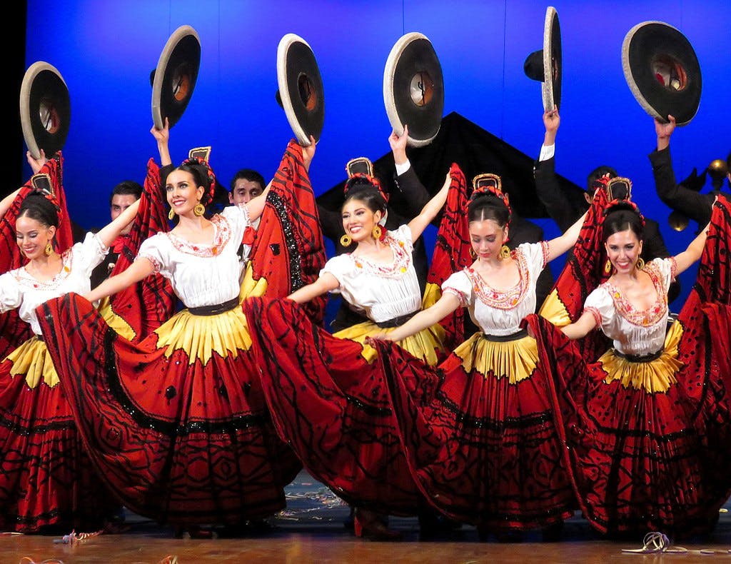 Tanečníci v barevných tradičních oděvech vystupují na jevišti, zvedají sukně a drží klobouky na modrém pozadí.